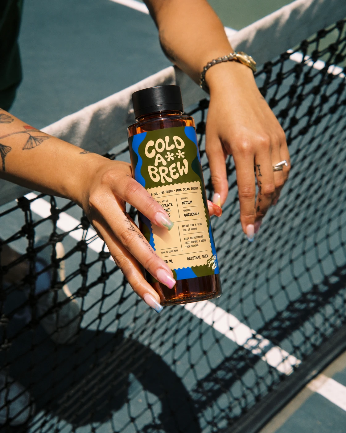 A hand holding a bottle of Cold A** Brew at the net of a court