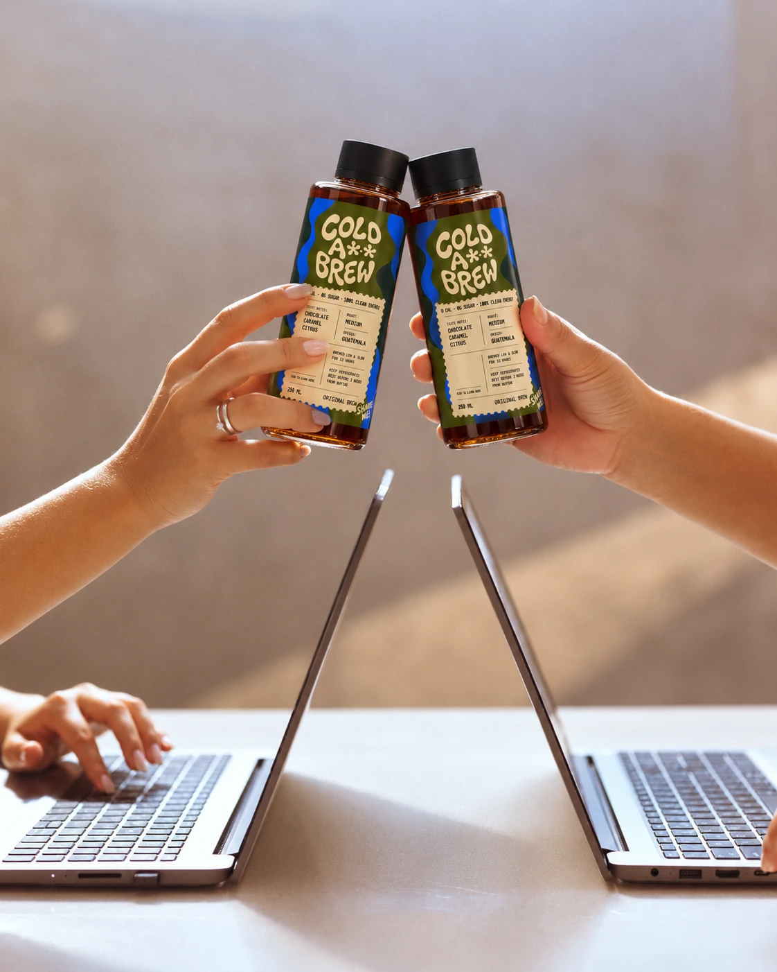 Two people toasting with Cold A** Brew bottles over their open laptops